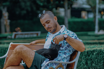 Young man touching the black cat in the park. Man with sun glasses touching the black cat on a bank in the park. Man loves the black cat outside.