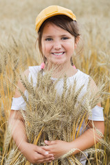 Cheerful girl in a yellow baseball cap with pigtails smiles and hugs ripe wheat in a field