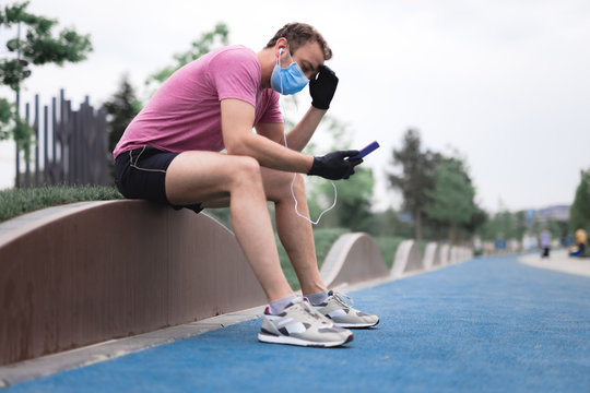 Sportsman With Medical Mask And Gloves, Smartphone And Earbuds Working Out, Jogging In Urban Surroundings. Exhausted Man From Jogging And Medical Mask Usage. Trouble With Breathing.