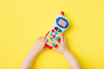 Baby hands playing with colorful toy of mobile phone on bright yellow table background. Closeup....