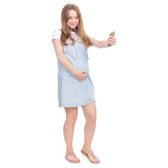 Pregnant woman in denim sundress with blue bag standing looking smiling, making selfie on smart phone on white background isolation