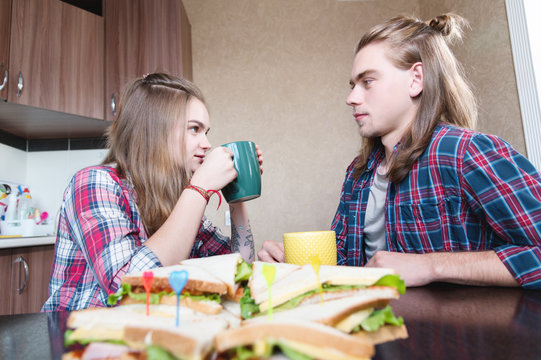 A Young Family Drinks Tea With Sandwiches In The Kitchen At The Dinner Table And Looks At Each Other. A Married Couple With Long Hair Chats Over Tea In The Home Dining Room