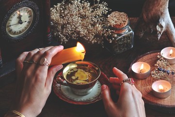 Wiccan witch wearing vintage jewelry holding yellow candle and pouring wax into a red gold vintage teacup as a divination. Reading Candle Wax - Carromancy, Ceroscopy among nature items dried flowers