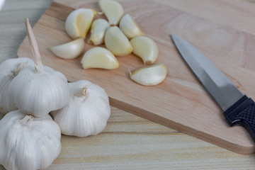 Peeled garlic slices with a knife stacked on a wood chopping board and with a pile of fresh garlic heads.