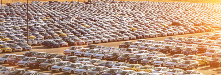 New cars parked in a distribution center on a sunset or sunrise and car factory buildings with pipes. Parking in the open air. © Eugene_Photo