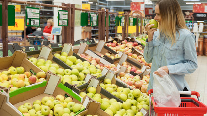 Portrait of a smiling attractive young woman shopper in the grocery shop during choosing and buying fresh apples at fruit department