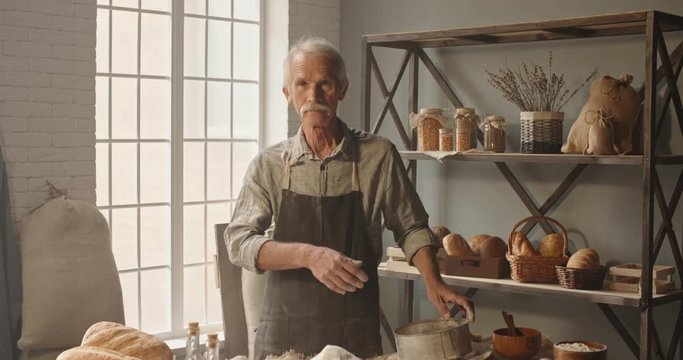 Joyful senior caucasian moustached man working as a professional baker at traditional bakery shop, looking at camera and positively smiling 4k footage