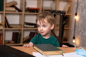 little boy sits at a table and looks at a laptop. near a book and a medical mask on the background of a bookcase. child looks at quarantine lessons online. serious look