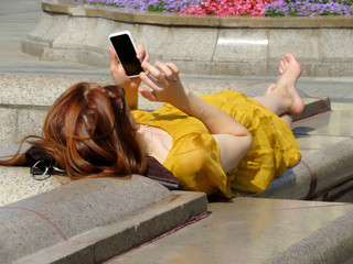 Barefoot woman in a summer dress lying with a smartphone in her hands in the city park. Concept of online communication, leisure outdoor, life after quarantine