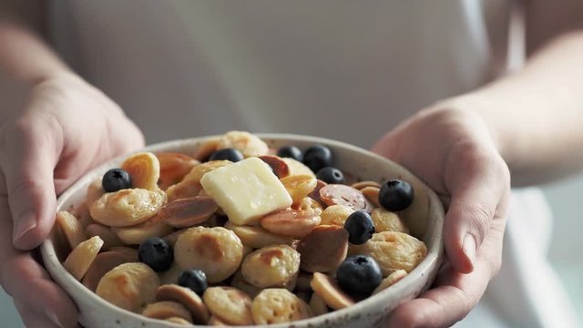 Bowl of mini pancakes in female hands. Trendy food - tiny panckaes or baby cereal pancakes served blueberries and butter. 4k footage. Camera shifting