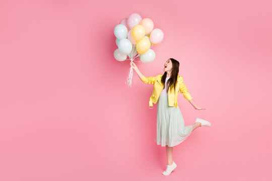 Full body profile photo of funny cheerful lady hold hands many air balloons fly up wind blowing wear yellow leather jacket green long skirt shoes isolated pastel pink color background