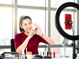 Asian beauty blogger or makeup artist woman review pink lipstick color show on her hand while record live through smartphone with selfie ring light for share on social media or channel in home studio