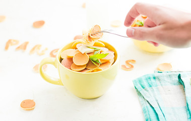 Mini pancake cereal in light green mug, white concrete background ready to serve on breakfast. New food trend. Copy space for text. Butter and mint leaves, milk, checkered aqua cloth. Hand with fork