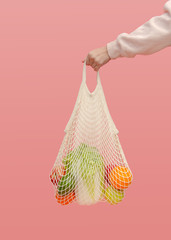 A woman's hand holds a mesh bag with vegetables and fruit on a pink background. The concept of zero waste and healthy food. Reusable shopping bag for going to the supermarket instead of plastic bags.