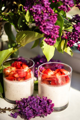 Vertical close-up picture of layered strawberry homemade italian creamy dessert panna cotta with fresh berries in two glasses with branch and bouquet of purple lilac. Organic sweet.