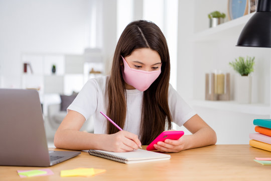 Photo of little pretty pupil small school lady laptop notebook online lesson sit desk use safety face mask write essay search telephone website distance quarantine study living room indoors