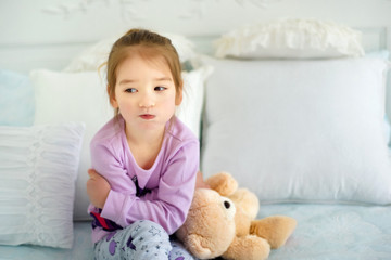 Portrait of small 3 years old sulking capricious girl sitting in bed in pajama with her toy bear. Emotional child. Kid in bad mood. Sad little girl in nightwear