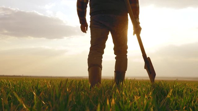 An agronomist man walks through a black plowed field at sunset. A farmer with a shovel in his hands and rubber boots.
