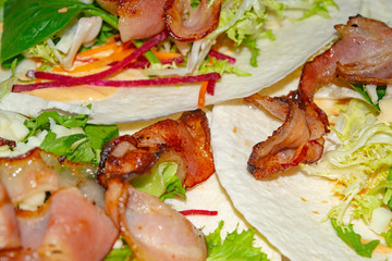 Leafy salad, sliced vegetables, sauce and fried bacon slices on pita bread are the basis for...