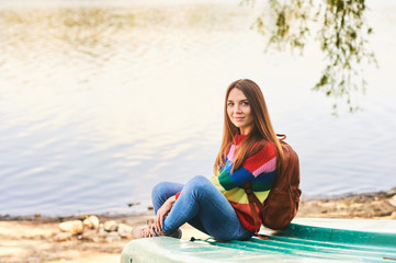 Fototapeta na wymiar Outdoor portrait of young beautiful woman relaxing by river on a nice warm day, wearing colorful pullover and backpack