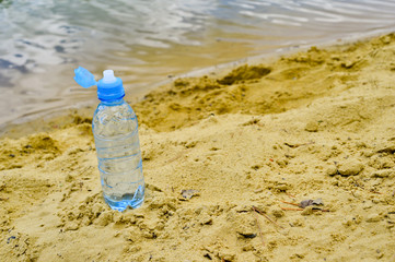 Fototapeta na wymiar An open plastic water bottle with a close-up stands on a sandy beach against the background of clear lake water.