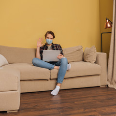 man in medical mask waving hand while having video call in living room