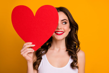 Photo of pretty cheerful lady hold big paper heart postcard hiding half facial expression interested look side empty space wear white casual singlet isolated yellow color background