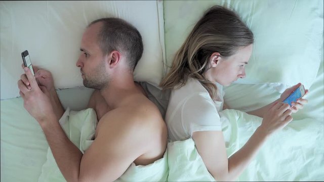 Couple lying in bed using mobile phone while ignoring each other. Man and woman addicted to smartphone social networks technology.