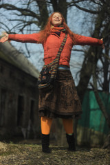 A happy red-haired young girl in a bright red jacket and pantyhose, in a skirt with floral patterns, poses against the backdrop of nature