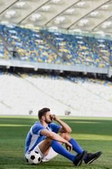 sad professional soccer player in blue and white uniform sitting with ball on football pitch at stadium © LIGHTFIELD STUDIOS