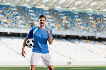 happy professional soccer player in blue and white uniform with ball showing yes gesture on...
