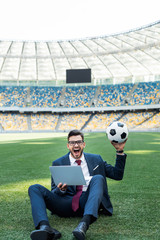 happy young businessman in suit with laptop and soccer ball sitting on football pitch at stadium,...