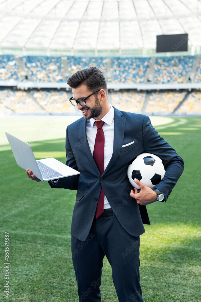 Wall mural smiling young businessman in suit with laptop and soccer ball at stadium, sports betting concept