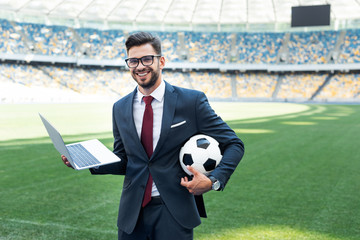 smiling young businessman in suit with laptop and soccer ball sitting on football pitch at stadium, sports betting concept