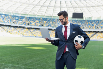 smiling young businessman in suit with laptop and soccer ball at stadium, sports betting concept