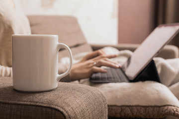 A white mug stands on the arm of a brown sofa against the blurry background of a girl's hands are typing on a laptop. Concept of distance work.