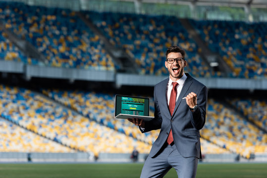 happy young businessman in suit holding laptop with sports betting website and showing yes gesture at stadium