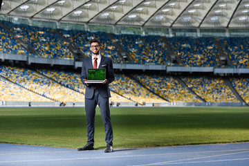 smiling young businessman in suit showing laptop with online trade website at stadium