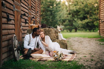 Boho style wedding. African groom and Caucasian bride are sitting on a plaid near the barn.