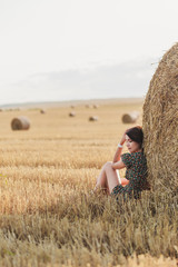Naklejka na ściany i meble A young beautiful girl in a dress is sitting by the hay in a field with haystacks