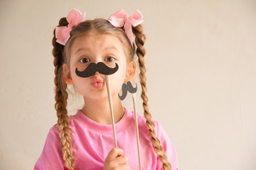 A little girl with pigtails is playing with a festive mustache.