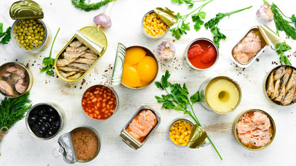 Food background in tin cans. Canned vegetables, beans, fish and fruits on a white wooden background. Top view.