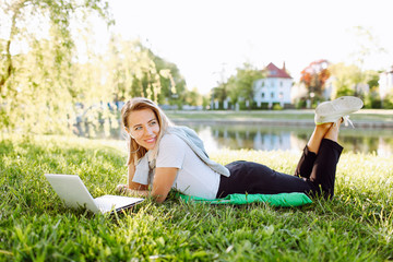 Girl working at the computer or having video chat lying on the grass in a park. Freelance, distance education concept.