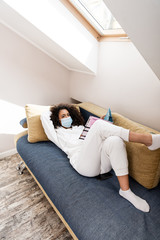 african american woman in medical mask lying on sofa and reading magazine