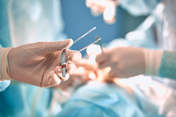 Doctor's hand holding tweezers in the operating room. Surgical instruments in the doctor s hand on the background of the operating room with doctors. The concept of medicine and surgery. Soft focus