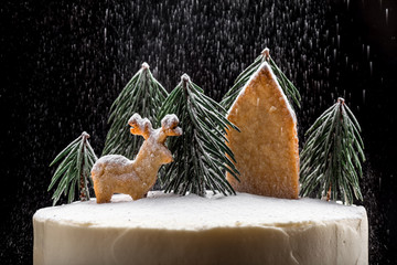 White winter rustic cake decorated with fir branches and cookies in the shape of a deer and the house sprinkled with powdered sugar, close-up on a dark background. 