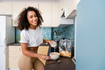 happy african american woman pouring milk in bowl with cornflakes