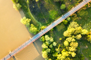 Aerial view of a narrow road bridge stretching over muddy wide river in green rural area.