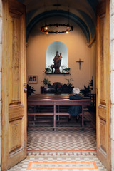 Old woman sitting alone on the pewage in catholic church and looking at Lord's table and the figure of Virgin Mary with baby Jesus on her hands, view through he doors