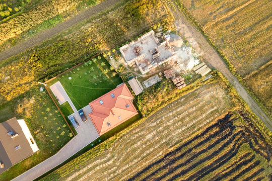 Top down aerial view of two private houses, one under construction with concrete foundament and brick walls and another finished with red tiled roof.